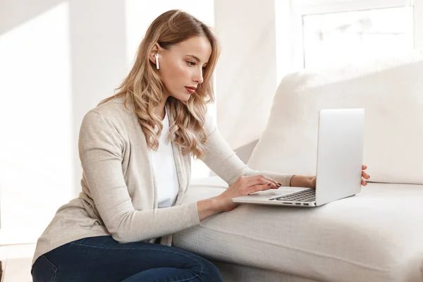 Bella donna bionda in posa seduta al chiuso a casa utilizzando il computer portatile . — Foto Stock