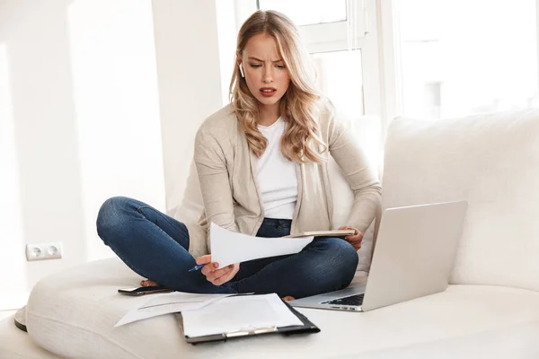 Confused blonde woman posing sitting indoors at home using laptop computer writing notes.