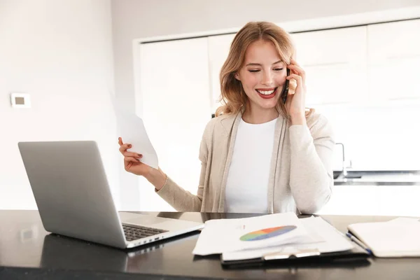 Beautiful blonde woman posing sitting indoors at home using laptop computer talking by mobile phone. — Stock Photo, Image