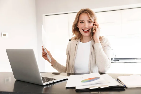 Hermosa mujer rubia posando sentada en casa usando computadora portátil hablando por teléfono móvil . — Foto de Stock