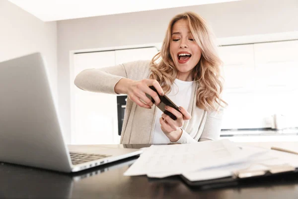 Femme blonde émotionnelle posant assis à l'intérieur à la maison en utilisant un ordinateur portable jouer à des jeux par téléphone mobile . — Photo