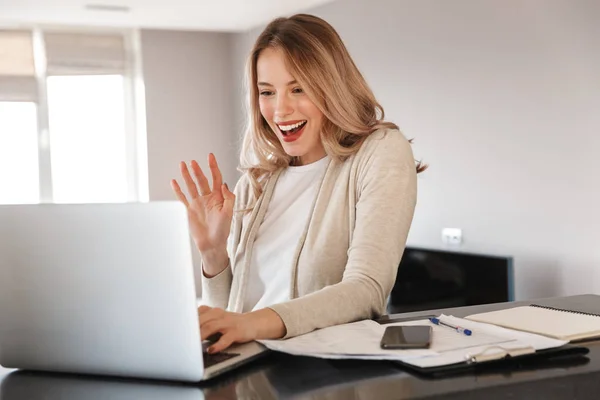 Beautiful blonde woman posing sitting indoors at home using laptop computer talking waving.