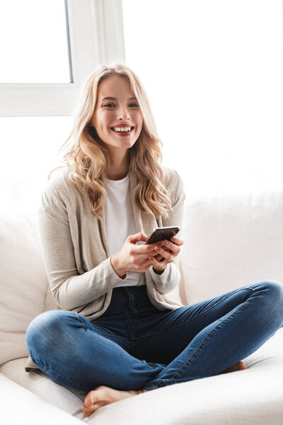 Beautiful blonde woman posing sitting indoors at home using mobile phone.