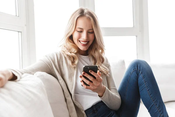 Beautiful blonde woman posing sitting indoors at home using mobile phone.