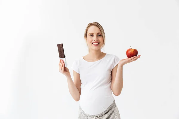 Krásná těhotná žena držící čokoládovou tyčinku a červené jablko — Stock fotografie