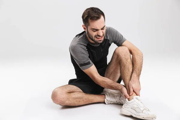 Young sports fitness man sitting on floor isolate over white wall background with painful feelings in leg. — Stock Photo, Image