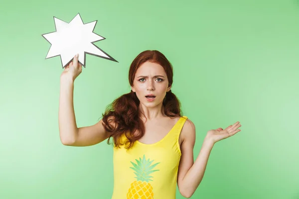 Shocked scared young redhead girl posing isolated over green wall background holding speech bubble. — Stock Photo, Image