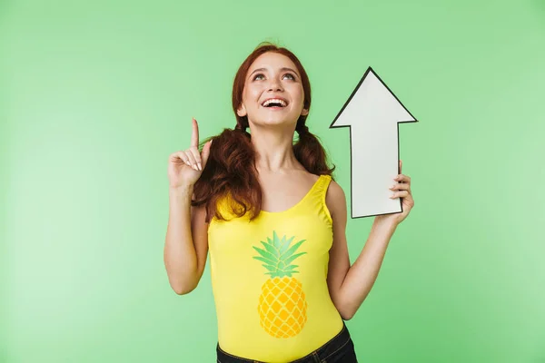 Hermosa joven pelirroja posando aislada sobre fondo de pared verde sosteniendo flecha . —  Fotos de Stock