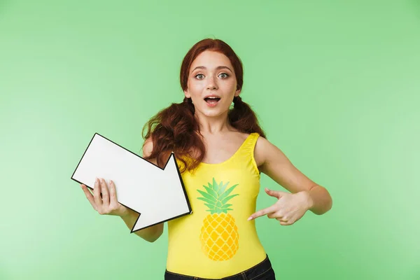 Beautiful young redhead girl posing isolated over green wall background holding arrow. — Stock Photo, Image