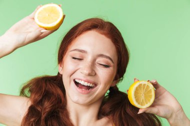 Young redhead girl posing isolated over green wall background with citrus vitamins fruits.