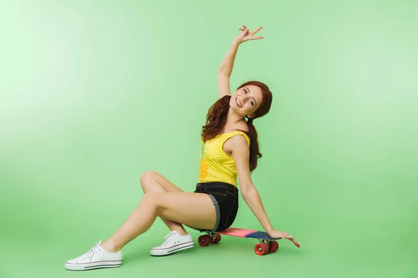 Lycklig ung rödhårig flicka poserar isolerade över grön vägg bakgrund med skateboard. — Stockfoto