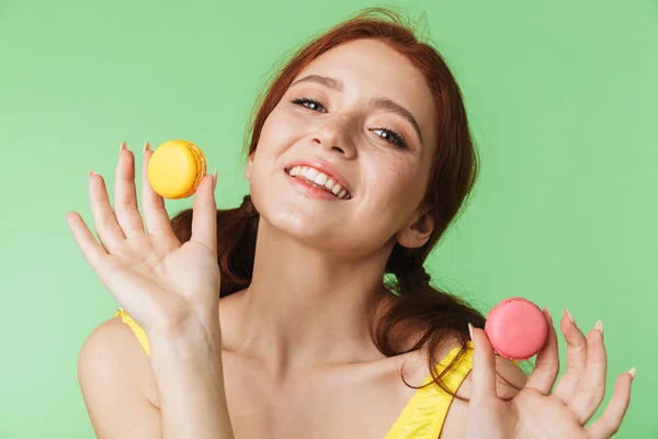 Redhead kız yeşil duvar arka plan üzerinde sweeties macaroons tutarak izole poz. — Stok fotoğraf