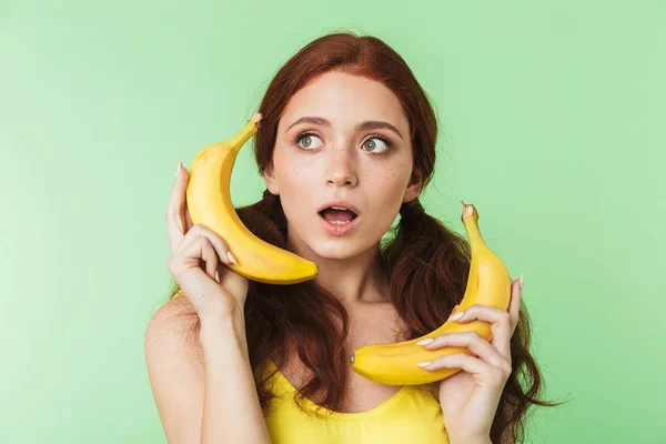 Upphetsad ung rödhårig flicka poserar isolerade över grön vägg bakgrund med bananer frukter. — Stockfoto