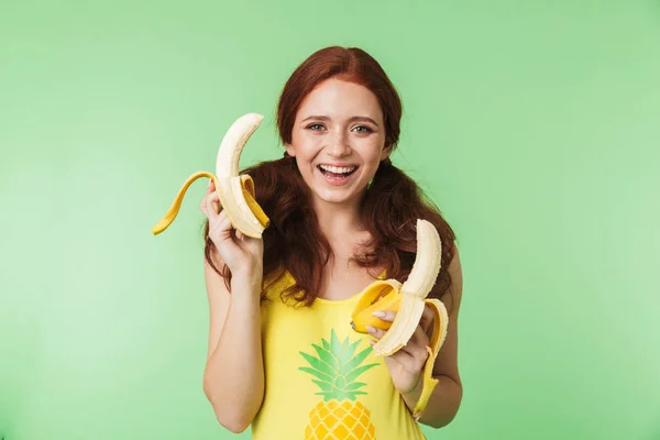 Excited young redhead girl posing isolated over green wall background with bananas fruits. — Stock Photo, Image
