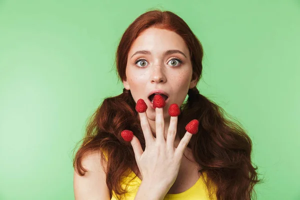 Schöne emotionale junge rothaarige Mädchen posiert isoliert über grüne Wand Hintergrund mit Himbeere. — Stockfoto