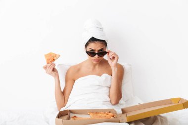 Kafasında havlu olan kadın battaniyenin altında yatıyor. Beyaz duvar arkasında izole edilmiş, güneş gözlüğü takıyor ve pizza yiyor..