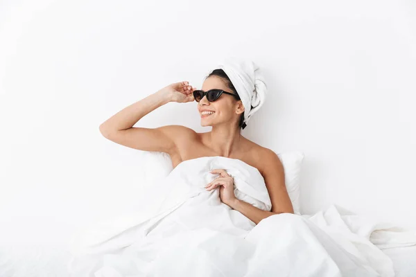 Bella donna incredibile con asciugamano sulla testa si trova nel letto sotto coperta isolata su sfondo bianco parete indossando occhiali da sole . — Foto Stock
