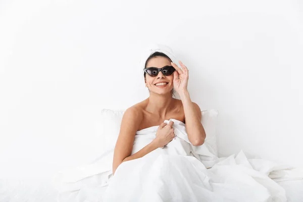 Bella donna incredibile con asciugamano sulla testa si trova nel letto sotto coperta isolata su sfondo bianco parete indossando occhiali da sole . — Foto Stock
