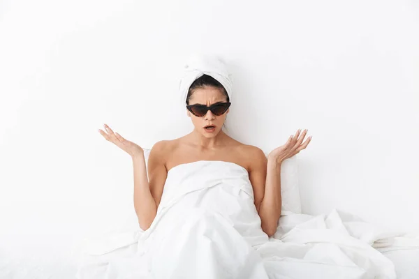Mulher chocada com toalha na cabeça encontra-se na cama sob cobertor isolado sobre fundo da parede branca usando óculos de sol . — Fotografia de Stock