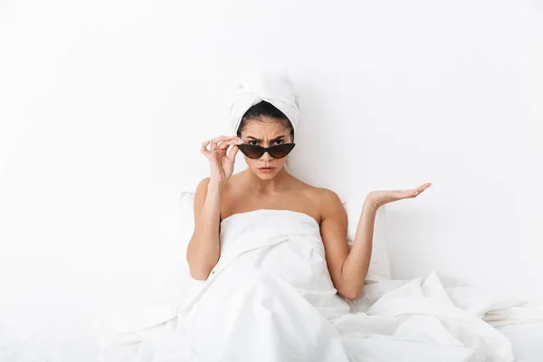 Geschokt vrouw met handdoek op hoofd ligt in bed onder deken geïsoleerd over witte muur achtergrond dragen zonnebril. — Stockfoto