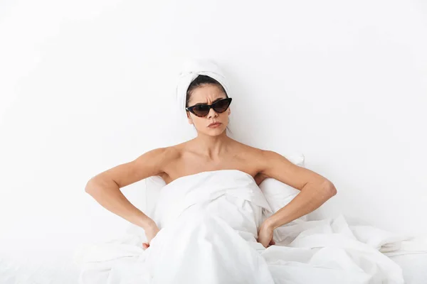 Hermosa mujer conmocionada con toalla en la cabeza se encuentra en la cama debajo de la manta aislada sobre fondo de pared blanca con gafas de sol . —  Fotos de Stock