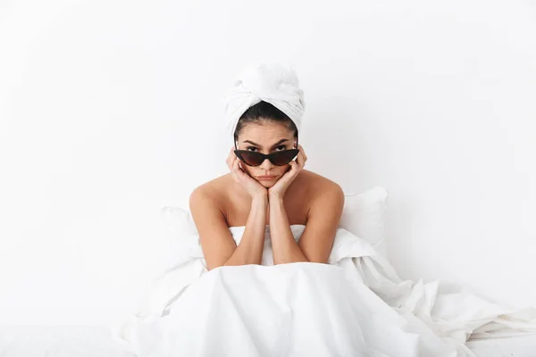 Donna con asciugamano sulla testa si trova nel letto sotto coperta isolato su sfondo bianco parete indossando occhiali da sole . — Foto Stock