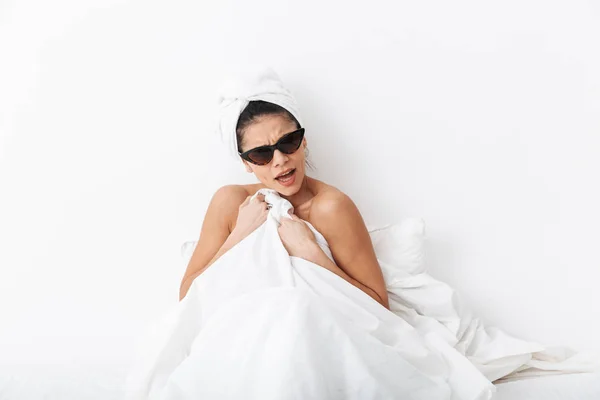 Mujer disgustada con toalla en la cabeza se encuentra en el cuerpo de la cama cubierta bajo manta aislada sobre fondo blanco de la pared usando gafas de sol . — Foto de Stock