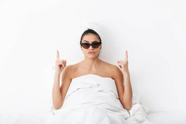 Женщина с полотенцем на голове лежит в постели покрывая тело под одеялом изолированы на белом фоне стены в солнечных очках указывая . — стоковое фото