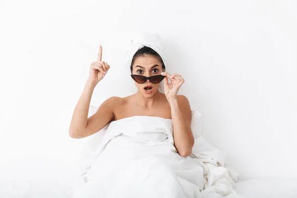 머리에 수건을 가진 흥분 한 여자는 선글라스를 입고 흰색 벽 배경 위에 고립 된 담요 아래 몸을 덮고 침대에 누워 있다.. — 스톡 사진