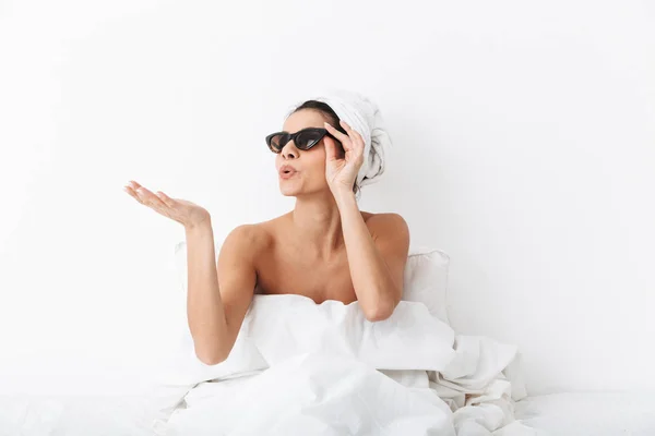 Mujer con toalla en la cabeza se encuentra en la cama bajo una manta aislada sobre fondo blanco de la pared con gafas de sol soplando besos . —  Fotos de Stock
