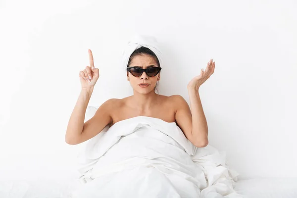 Woman with towel on head lies in bed covering body under blanket isolated over white wall background wearing sunglasses pointing.