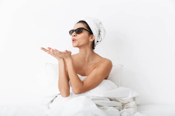 Mujer con toalla en la cabeza se encuentra en la cama bajo una manta aislada sobre fondo blanco de la pared con gafas de sol soplando besos . —  Fotos de Stock