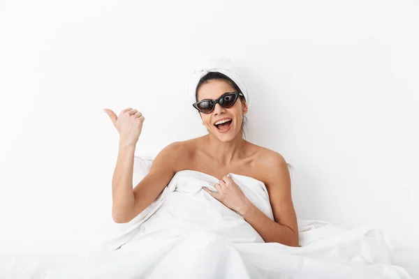 머리에 수건을 가진 행복한 여자는 선글라스를 입고 흰색 벽 배경 위에 고립 된 담요 아래 침대에 누워 있습니다.. — 스톡 사진