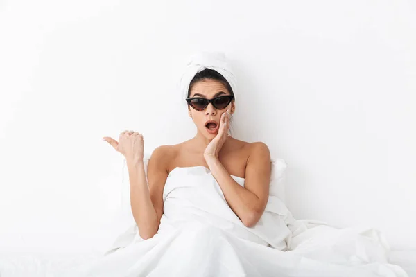 Krásná šokovaná žena s ručníkem na hlavě leží v posteli pod přikrývkou izolovanou na bílém pozadí, nosí sluneční brýle ukazující. — Stock fotografie