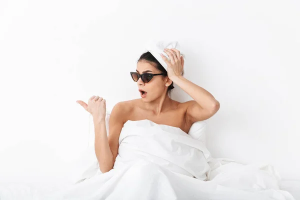 Hermosa mujer conmocionada con toalla en la cabeza se encuentra en la cama debajo de la manta aislada sobre fondo de pared blanca con gafas de sol apuntando . —  Fotos de Stock