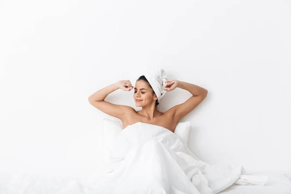Bella donna incredibile con asciugamano sulla testa si trova nel letto sotto coperta isolato su sfondo bianco parete . — Foto Stock