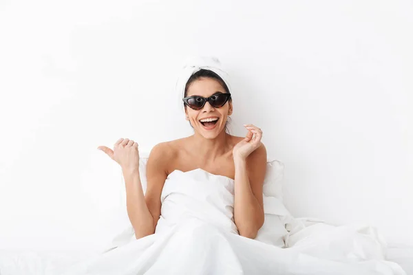 Szczęśliwa kobieta z ręcznikiem na głowie leży w łóżku pod kocem na białym tle ściany noszenie okularów przeciwsłonecznych wskazując. — Zdjęcie stockowe