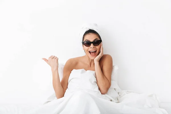 Donna felice con asciugamano sulla testa si trova nel letto sotto coperta isolato su sfondo bianco parete indossando occhiali da sole che puntano . — Foto Stock