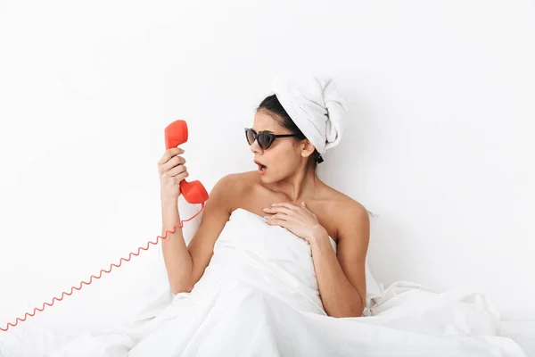 Mujer sorprendida descontenta con toalla en la cabeza se encuentra en la cama debajo de la manta aislada sobre fondo blanco de la pared usando gafas de sol hablando por teléfono . —  Fotos de Stock