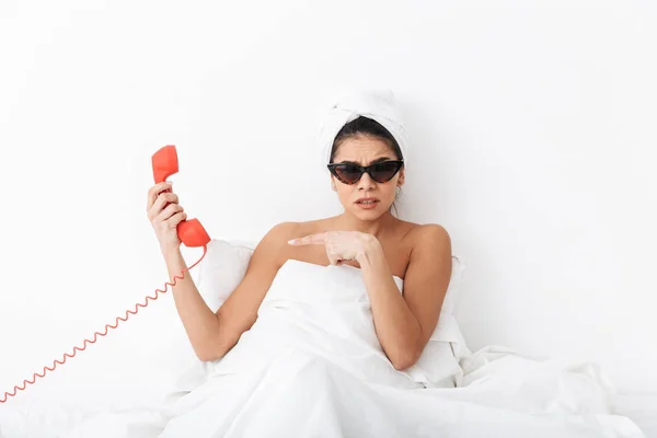Mujer sorprendida descontenta con toalla en la cabeza se encuentra en la cama debajo de la manta aislada sobre fondo blanco de la pared usando gafas de sol hablando por teléfono . — Foto de Stock