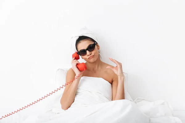 Başında havlu ile rahatsız kadın telefonla konuşurken güneş gözlüğü giyen beyaz duvar arka plan üzerinde izole battaniye altında yatakta yatıyor. — Stok fotoğraf
