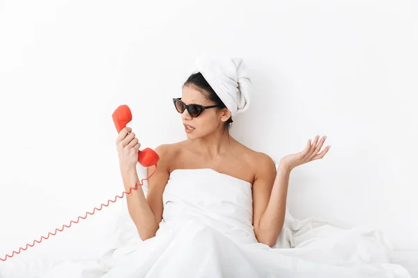 Nelibě šokovaná žena s ručníkem na hlavě leží v posteli pod přikrývkou izolovanou na bílém pozadí, nosí sluneční brýle, mluvící telefonem. — Stock fotografie