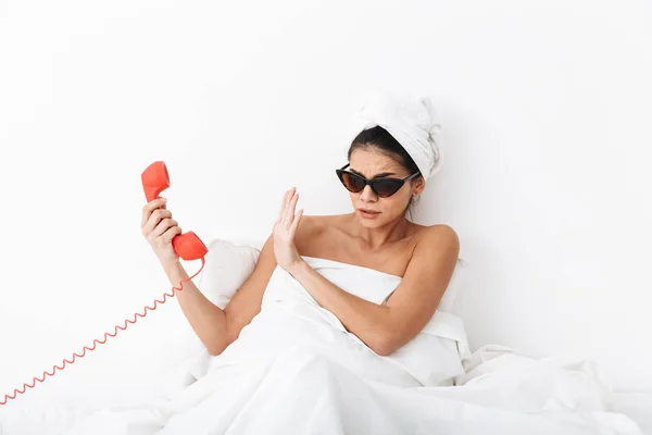 Mujer sorprendida descontenta con toalla en la cabeza se encuentra en la cama debajo de la manta aislada sobre fondo blanco de la pared usando gafas de sol hablando por teléfono . —  Fotos de Stock