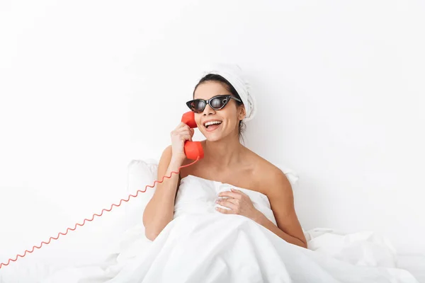 Mulher emocional com toalha na cabeça encontra-se na cama sob cobertor isolado sobre fundo da parede branca usando óculos de sol falando por telefone . — Fotografia de Stock