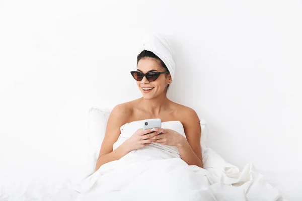Емоційна жінка з рушником на голові лежить в ліжку під ковдрою ізольована на білому тлі стіни в сонцезахисних окулярах, спілкуючись по телефону . — стокове фото