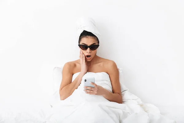 Hermosa mujer emocional sorprendida con toalla en la cabeza se encuentra en la cama debajo de la manta aislada sobre fondo blanco de la pared usando gafas de sol charlando por teléfono . — Foto de Stock