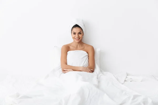 Hermosa mujer increíble con toalla en la cabeza se encuentra en la cama debajo de la manta aislada sobre fondo de pared blanca . — Foto de Stock