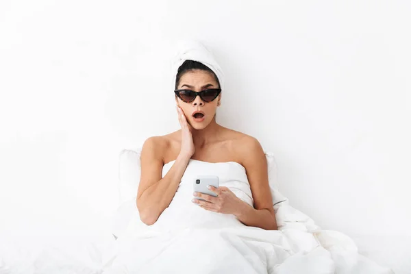 頭にタオルを持つ美しい驚いた感情的な女性は、電話でおしゃべりサングラスを着て白い壁の背景の上に隔離された毛布の下にベッドに横たわっています. — ストック写真