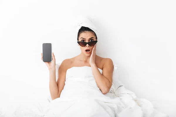 頭にタオルを持つ驚いた感情的な女性は、携帯電話の表示を示すサングラスを着て白い壁の背景の上に隔離された毛布の下にベッドに横たわっています. — ストック写真