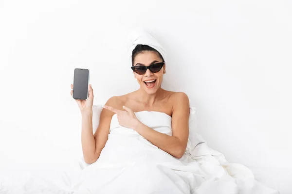 Удивительная эмоциональная женщина с полотенцем на голове лежит в постели под одеялом изолированы на белом фоне стены в солнечных очках, показывающих дисплей телефона . — стоковое фото